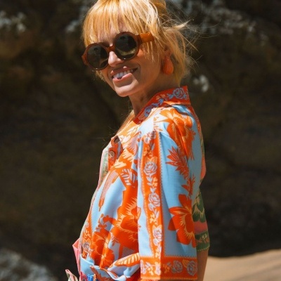 Blusa floral azul claro e laranja com colarinho e botões, óculos de sol redondos castanhos em mulher na praia