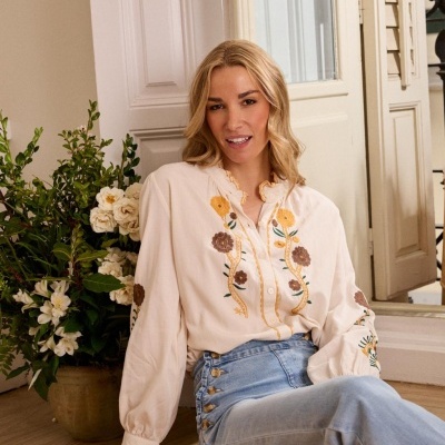 Camisa feminina branca com bordados florais e calças de ganga azul claro sentada no chão junto a plantas.