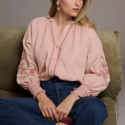 Mulher sentada numa poltrona verde com blusa rosa bordada e calças de ganga azuis.