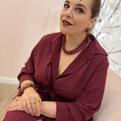 Mulher com vestido bordeaux e joias em pedras vermelhas sentada em ambiente claro com flores.