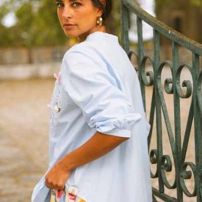 Camisa azul clara bordada e saia colorida junto a grade de ferro verde