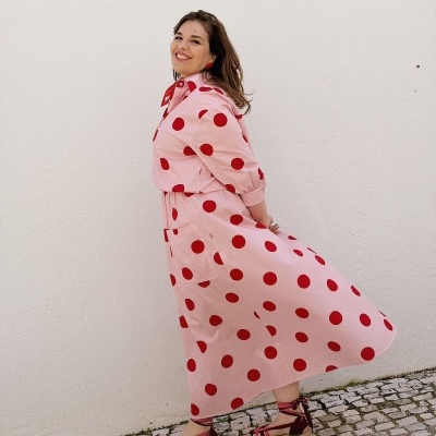 Vestido rosa comprido com bolinhas vermelhas usado por mulher com sandálias vermelhas junto a parede branca