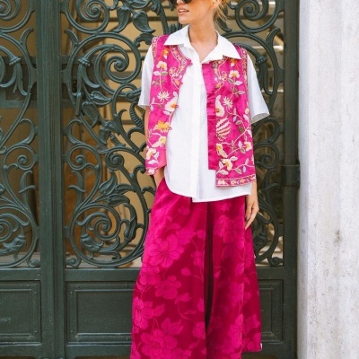 Mulher com conjunto rosa bordado, camisa branca, calças largas com padrão floral e sapatos prateados, com óculos de sol, em porta de ferro trabalhado