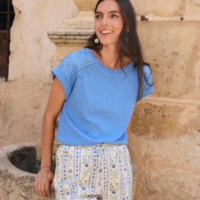 Mulher com t-shirt azul renda nos ombros e calças florais ao lado de fonte em pedra.