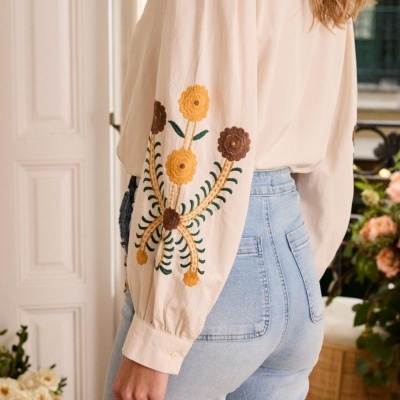 Blusa creme com bordados florais nas mangas e jeans azul claro num ambiente interior decorado com flores