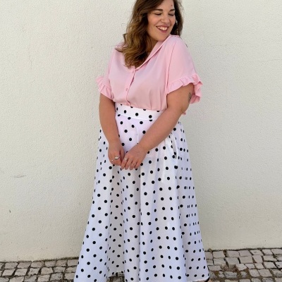 Mulher a sorrir com blusa rosa e saia branca de bolinhas pretas numa calçada