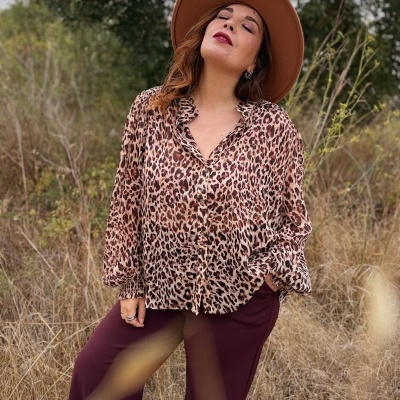 Mulher com camisa animal print, calças bordeaux e chapéu castanho em cenário natural com vegetação seca
