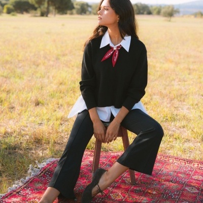 Mulher sentada na relva com roupa preta e branca e lenço vermelho, sobre tapete vermelho com padrão.
