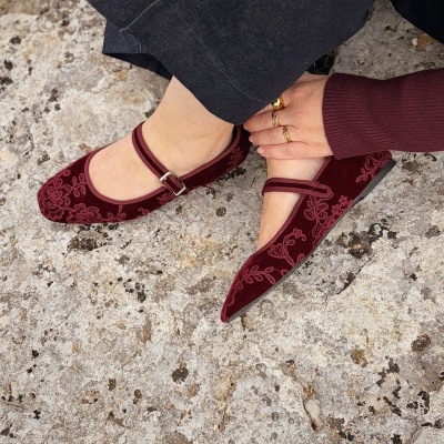Sapatos Mary Jane em veludo bordô com bordados florais, fivela prateada, calças pretas, superfície de pedra.