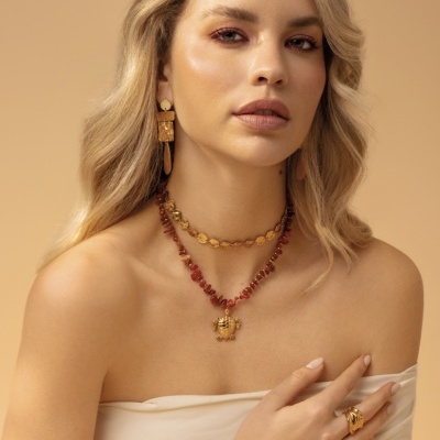 Mulher com joias douradas e vermelhas, incluindo colares, brincos, anel e pulseira, em fundo bege.