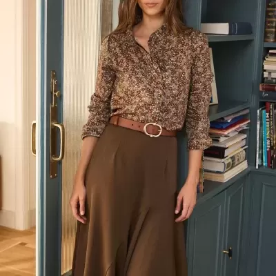 Mulher com camisa floral castanha e saia midi castanha junto a estante azul com livros