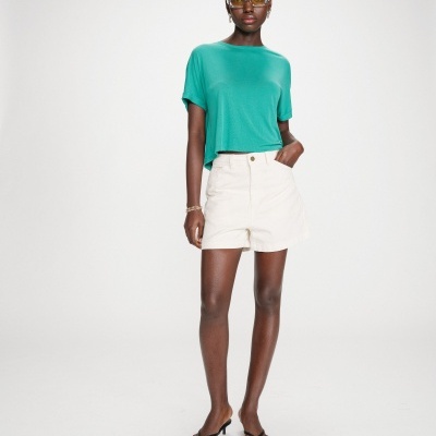 Mulher com t-shirt verde, calções brancos e sandálias pretas, usando óculos de sol com lentes amarelas.