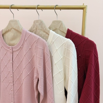 Três camisolas de malha com padrão de losangos e pérolas nas cores rosa, branca e bordo penduradas em cabides de tecido bege