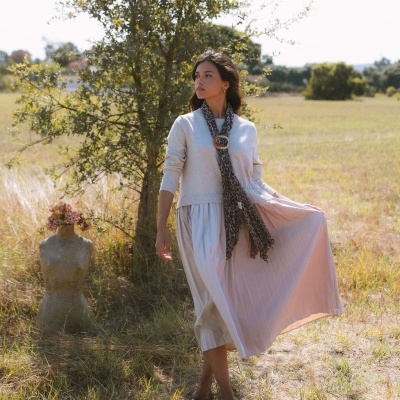 Mulher com vestido branco e lenço animal print num campo com grama seca e árvore, ao lado de manequim com flores