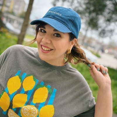 Mulher sorridente com boné azul e t-shirt cinza com limões amarelos e texto em rosa ao ar livre