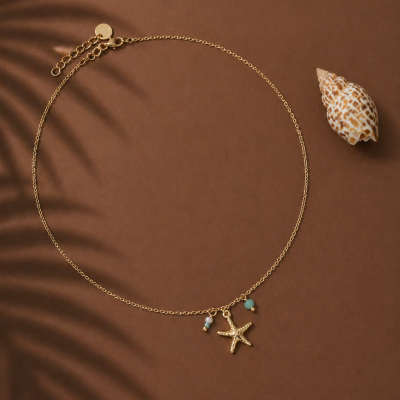 Colar dourado com pingente de estrela do mar e pedras azuis numa superfície castanha com sombra de folha e concha