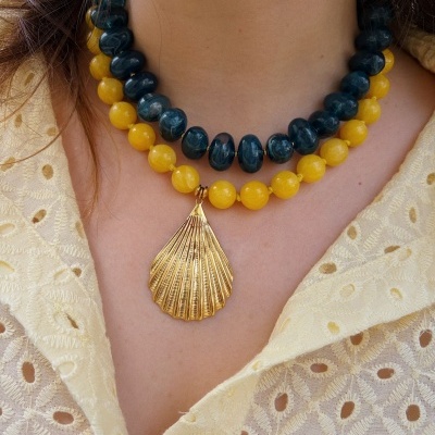 Colares de contas azul escuro e amarelo com pendente dourado em forma de concha, usados sobre camisa amarela bordada.
