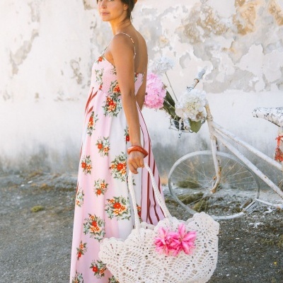 Mulher com vestido floral e mala branca em crochet com flor rosa, junto a bicicleta branca com flores, parede velha ao fundo.