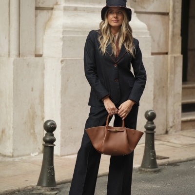 Mulher com conjunto preto, chapéu preto, mala castanha, botas pretas, em ambiente urbano com parede de pedra clara ao fundo