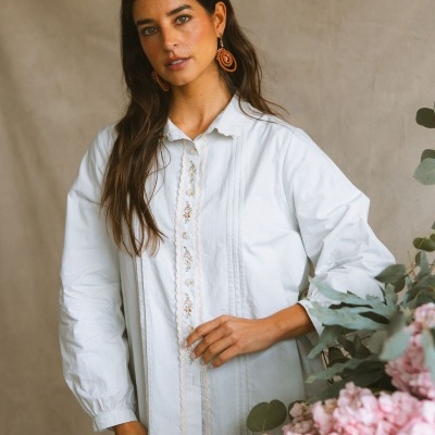 Blusa branca com bordado floral e renda, usada por mulher com brincos castanhos perto de flores rosas.