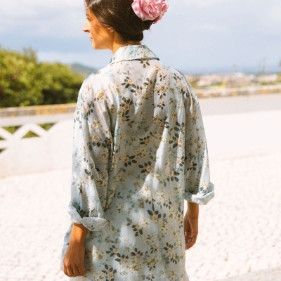 Mulher de costas com camisa azul clara de padrão floral e flor rosa no cabelo ao ar livre.
