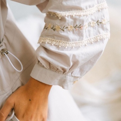 Manga de blusa branca com bordado floral e renda, punho franzido, mão com tatuagem pequena.