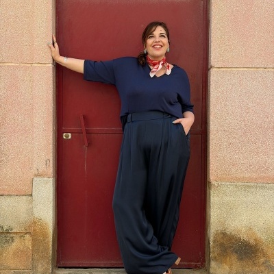 Mulher com blusa azul escura, calças largas azuis, lenço vermelho e sandálias castanhas junto a porta vermelha