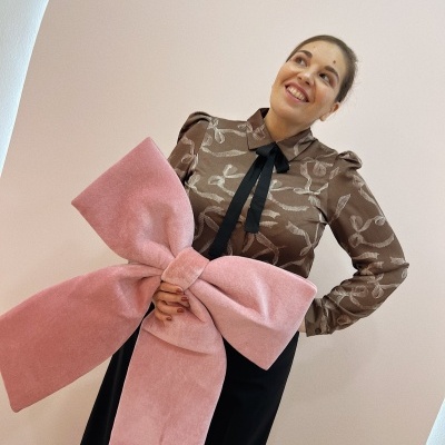 Mulher com blusa castanha estampada e laço preto, calças pretas e grande laço rosa aveludado