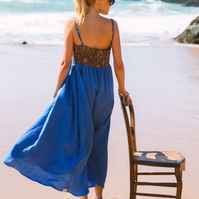 Mulher de costas na praia com vestido longo azul e parte superior floral castanha, segurando cadeira de madeira.