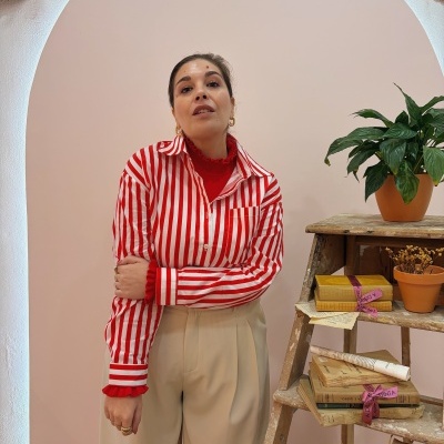 Mulher com camisa de riscas vermelhas e brancas e calças bege junto a escada com plantas e livros