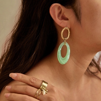 Brincos dourados e verdes em formato oval com anéis dourados na mão