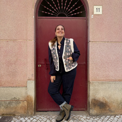 Mulher vestindo colete floral com bordas brancas, camisa azul, calças de ganga e botas verdes em frente a porta vermelha com número 11