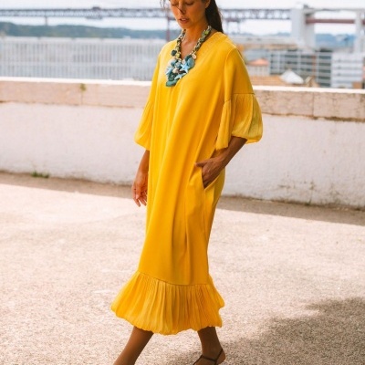 Vestido amarelo midi com mangas e barra plissadas usado com sandálias e colar em ambiente urbano