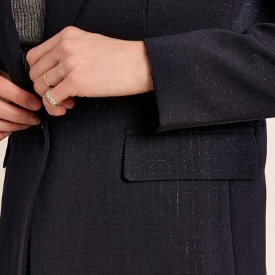 Close-up de blazer feminino tecido escuro com brilho e bolso, com pessoa a abotoar e camisola cinzenta por baixo.