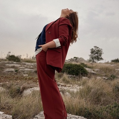 Mulher com fato vermelho vinho, camisa branca e colete azul em ambiente exterior rochoso