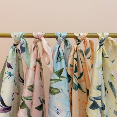 Seis lenços de seda com padrões florais em cores pastel pendurados numa barra dourada.