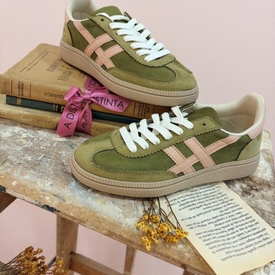 Sapatilhas verdes com detalhes rosa e atacadores brancos sobre mesa rústica com livros e flores secas.