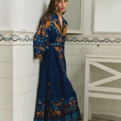 Mulher com vestido azul escuro estampado encostada a uma parede de azulejos brancos.