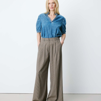 Mulher com blusa azul de ganga, calças largas xadrez castanho e sapatos castanhos em fundo branco