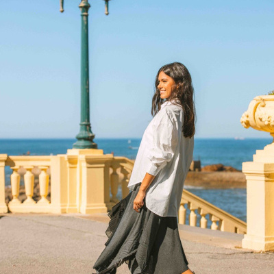 Mulher com camisa branca, saia cinzenta e botas pretas ao ar livre junto ao mar