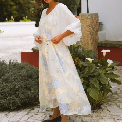 Vestido longo floral branco com mangas largas e cinto, sapatos pretos, mulher em pavimento de pedra ao ar livre