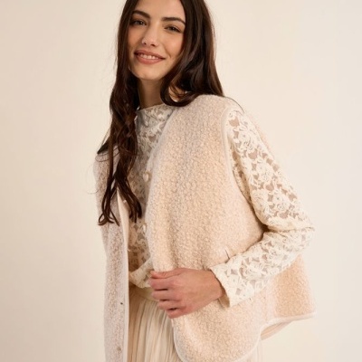 Mulher com colete bege peludo, blusa rendada branca e saia branca fluida