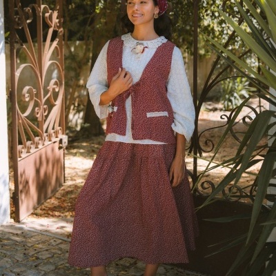 Mulher com colete e saia vermelhos com padrão floral, blusa branca com gola de renda, lenço na cabeça e sapatilhas castanhas em ambiente exterior.