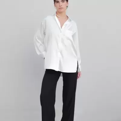 Mulher com camisa branca de tecido acetinado, calças pretas e sapatos xadrez em fundo cinzento.