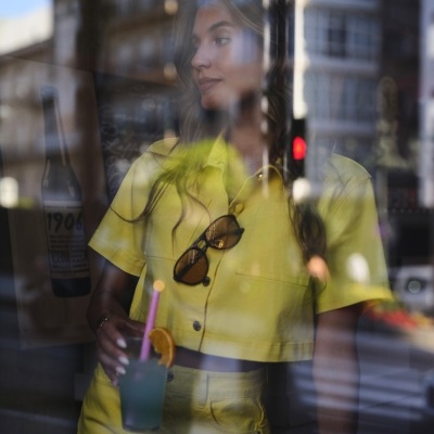 Mulher com roupa amarela, segurando bebida com canudo rosa, vista através de janela com reflexos urbanos