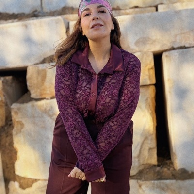 Mulher com lenço rosa, blusa de renda roxa e calças roxas em fundo de pedras