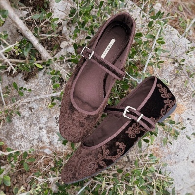 sapatos Mary Jane castanhos em veludo com bordados florais sobre chão de pedra e plantas