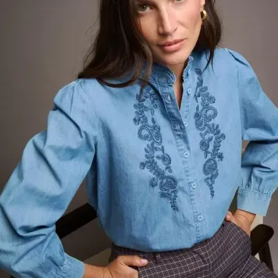 Blusa azul clara com bordado floral e calças xadrez castanhas usadas por mulher sentada