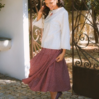 Mulher com blusa branca de manga longa e saia vermelha florida, lenço vermelho na cabeça e sapatos castanhos, posando junto a portão de ferro e parede branca