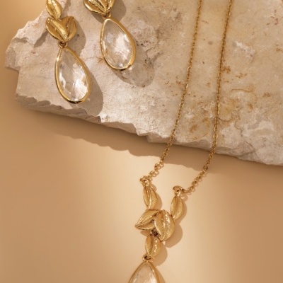 Conjunto de brincos e colar dourados com folhas e gotas transparentes sobre pedra clara